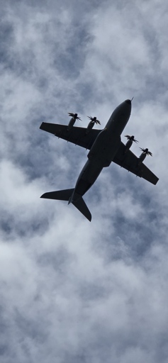 A large 4-engine turboprop military transport plane viewed from below, directly above camera at a steep angle, with several layers of clouds in the background